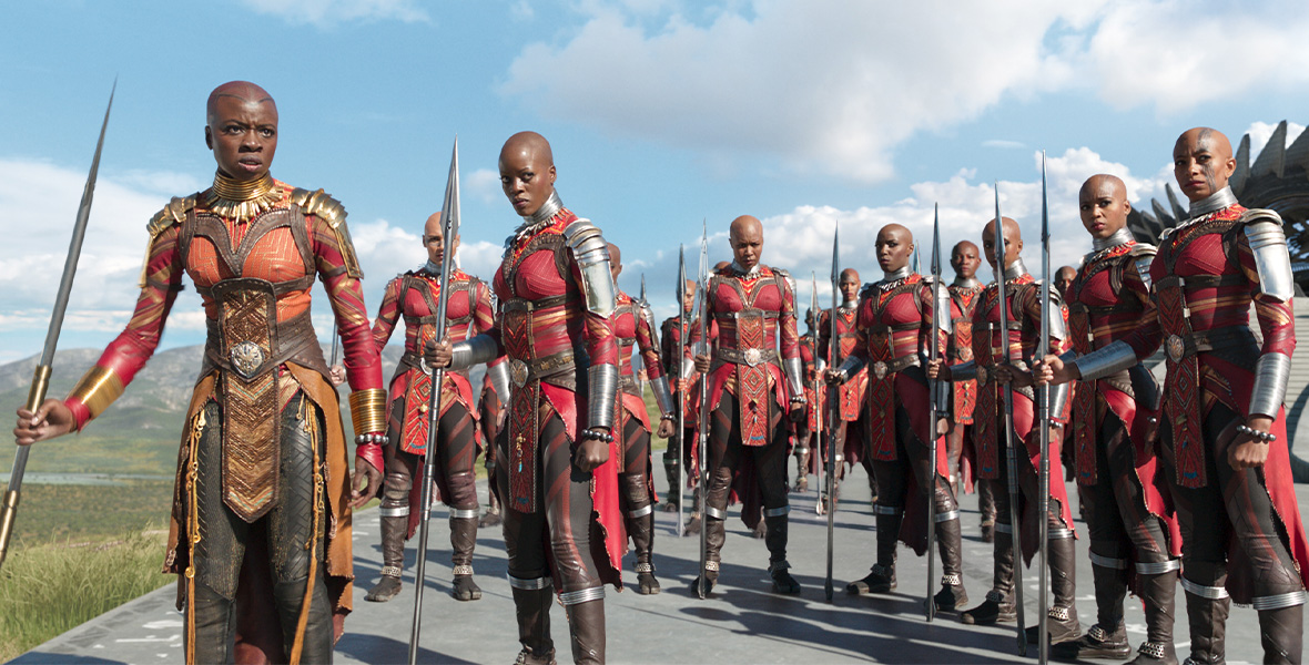 From the left Actor Danai Gurira portrays General Okoye from the Dora Milaje. Gurira and the Dora Milaje wear red and gold uniforms with intricate embroidery, brown leather pants, and brown leather boots. They hold tall metal spears in their left hand. The sky is blue with fluffy white clouds above them.