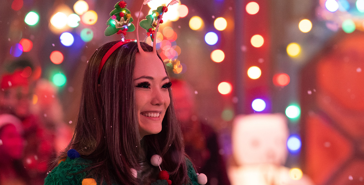 In a scene from Marvel Studios’ The Guardians of the Galaxy: Holiday Special, actor Pom Klementieff portrays Mantis. She wears a green knit sweater, red headband with two tiny Christmas trees, and a necklace with red, white, and green pompom balls. Out of focus in the background are colorful Christmas-themed lights and people wearing Christmas-themed hats and sweaters.
