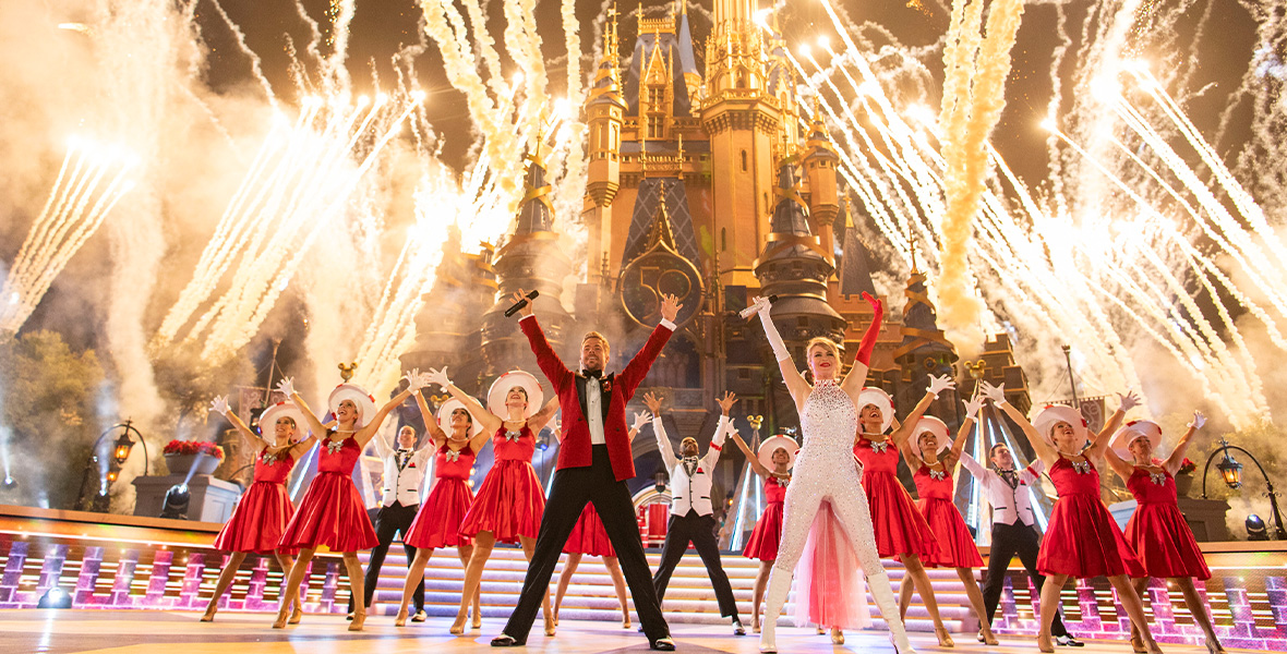 In a scene from The Wonderful World of Disney: Magical Holiday Celebration, performers Derek Hough and Julianne Hough stand center stage, with their arms extended up into the air, in front of Cinderella’s Castle at Walt Disney World Resort. Fireworks soar into the sky and pyrotechnics deploy around them. Behind Derek and Julianne stand a group of dancers wearing red and white outfits. Derek wears a cardinal velvet suit jacket with black lapels and black pants. Julianne wears a white sequin body suit with one white opera glove and one red opera glove.