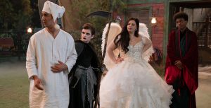 In a scene from BUNK’D: Learning the Robes, actors Trevor Tordjman, Shiloh Verrico, Miranda May, and Israel Johnson stand side by side and look to their right. Tordjman portrays Parker and wears a long, white night shirt with a matching cap. Verrico portrays Winnie and wears an all-black ensemble resembling the Grim Reaper and holds a clever in her left hand. May portrays Lou and wears a white ballgown with a beaded bodice and white angel wings. Johnson portrays Noah and wears a long red and black velvet cloak with a black robe underneath. Behind the actors is a rustic cabin with green shutters and pine trees.