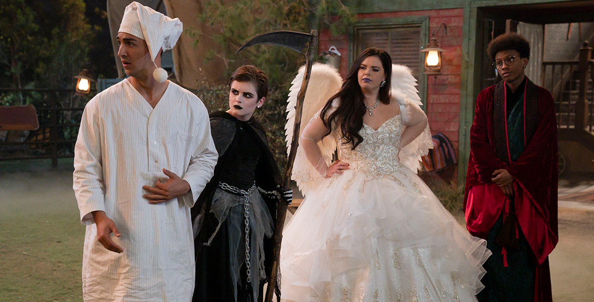 In a scene from BUNK’D: Learning the Robes, actors Trevor Tordjman, Shiloh Verrico, Miranda May, and Israel Johnson stand side by side and look to their right. Tordjman portrays Parker and wears a long, white night shirt with a matching cap. Verrico portrays Winnie and wears an all-black ensemble resembling the Grim Reaper and holds a clever in her left hand. May portrays Lou and wears a white ballgown with a beaded bodice and white angel wings. Johnson portrays Noah and wears a long red and black velvet cloak with a black robe underneath. Behind the actors is a rustic cabin with green shutters and pine trees.
