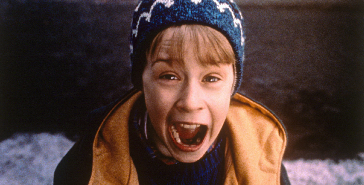 In a scene from the film Home Alone, actor Macaulay Culkin portrays Kevin McCallister and looks up and screams. In the close-up shot, Culkin wears a blue knitted beanie with a white design, a blue sweater, and a dark blue jacket with a tan fabric inside. Behind Culkin are black asphalt and snow-covered sidewalks.