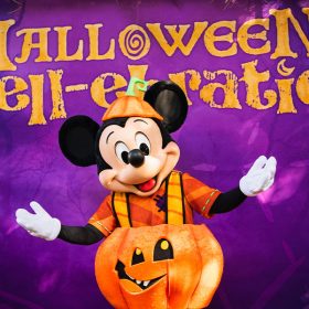 Mickey Mouse posing in front of a purple backdrop with “Halloween Spell-ebration” in yellow letters. Mickey is wearing his pumpkin costume that debuted at Disneyland Resort—a pumpkin hat in orange and green, an orange striped shirt, a pumpkin around his waist with orange suspenders, and purple striped pants.