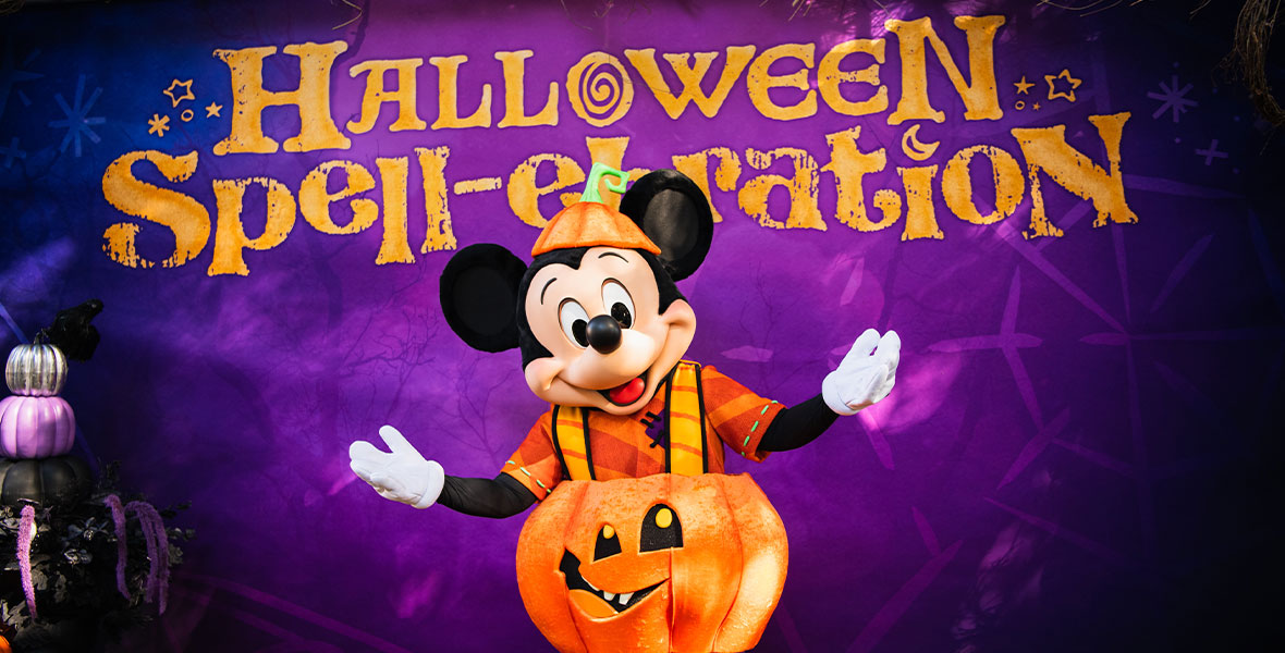 Mickey Mouse posing in front of a purple backdrop with “Halloween Spell-ebration” in yellow letters. Mickey is wearing his pumpkin costume that debuted at Disneyland Resort—a pumpkin hat in orange and green, an orange striped shirt, a pumpkin around his waist with orange suspenders, and purple striped pants.