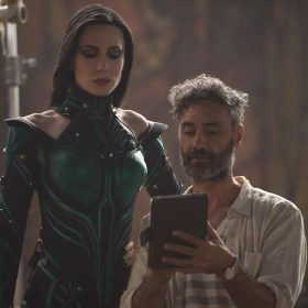 On the set of Thor: Ragnarok, actress Cate Blanchett towers over director Taika Waititi as they both watch something on an iPad together. Waititi is in casual clothes, while Blanchett is in full costume and makeup as her character, Hela, the goddess of death.