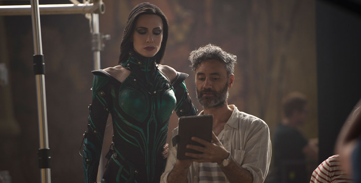On the set of Thor: Ragnarok, actress Cate Blanchett towers over director Taika Waititi as they both watch something on an iPad together. Waititi is in casual clothes, while Blanchett is in full costume and makeup as her character, Hela, the goddess of death.
