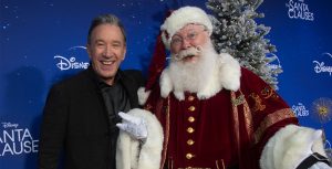 Tim Allen and Santa Claus pose together with big smiles in front of a snowy Christmas tree and a dark blue backdrop with Disney+ and The Santa Clauses branding.