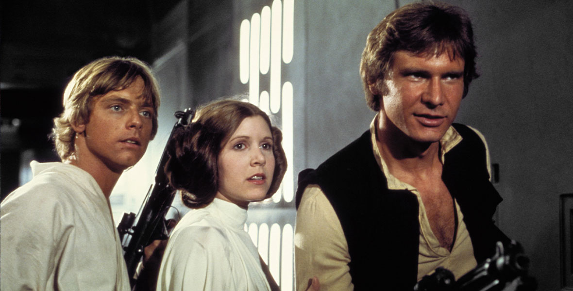 In a still from 1977’s Star Wars: A New Hope, Luke Skywalker (Disney Legend Mark Hamill), Princess Leia (Disney Legend Carrie Fisher), and Han Solo (Harrison Ford) are standing together looking off-camera to the right. Han is holding a blaster-type weapon in his hand and pointing it at someone; Luke is also holding a blaster but is pointing it upwards.