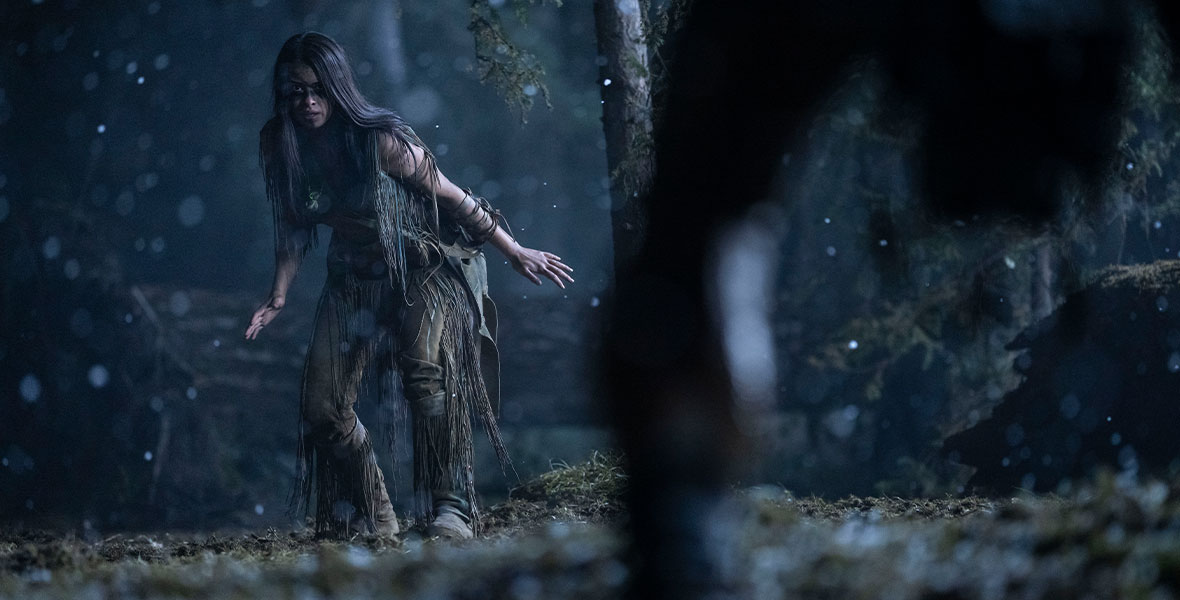 In a still from 2022’s Prey, Naru (Amber Midthunder) is in a forest and is looking at someone whose legs we see to the right of the image. It appears to be raining. Naru has long dark hair and her arms are outstretched in a way that makes her look ready to do battle.