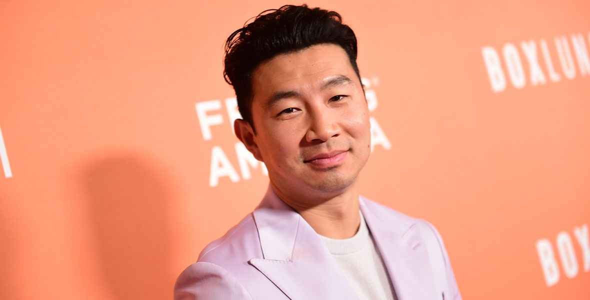Actor Simu Liu stands against a bright orange step-and-repeat and looks directly at the camera. He is wearing a lavender suit jacket with a white wool sweater underneath.
