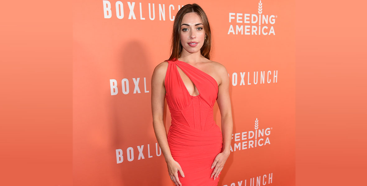 Isabella Ward of West Side Story stands on the red carpet against an orange step-and-repeat featuring the BoxLunch and Feeding America logos. She has long brown hair that sits straight behind her shoulders and she is wearing an ankle-length, one-shouldered red dress.