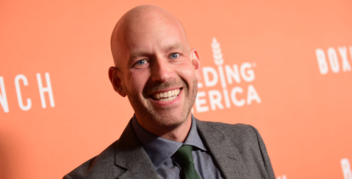Bret Iwan smiles at the camera in front of an orange step-and-repeat. He is wearing a gray suit jacket, gray button up shirt, and forest green tie.
