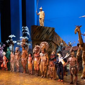 In a still from The Lion King on Broadway’s 25th anniversary performance on November 13, 2022, the company is taking their curtain call at the end of the show; they’re holding hands and looking out at the audience, smiling. Almost everyone is standing in a line downstage towards the edge of the physical stage, while one cast member up on “Pride Rock,” holding up a puppet of a bird. The backdrop behind the cast is blue. Photo by Chrystofer Davis.