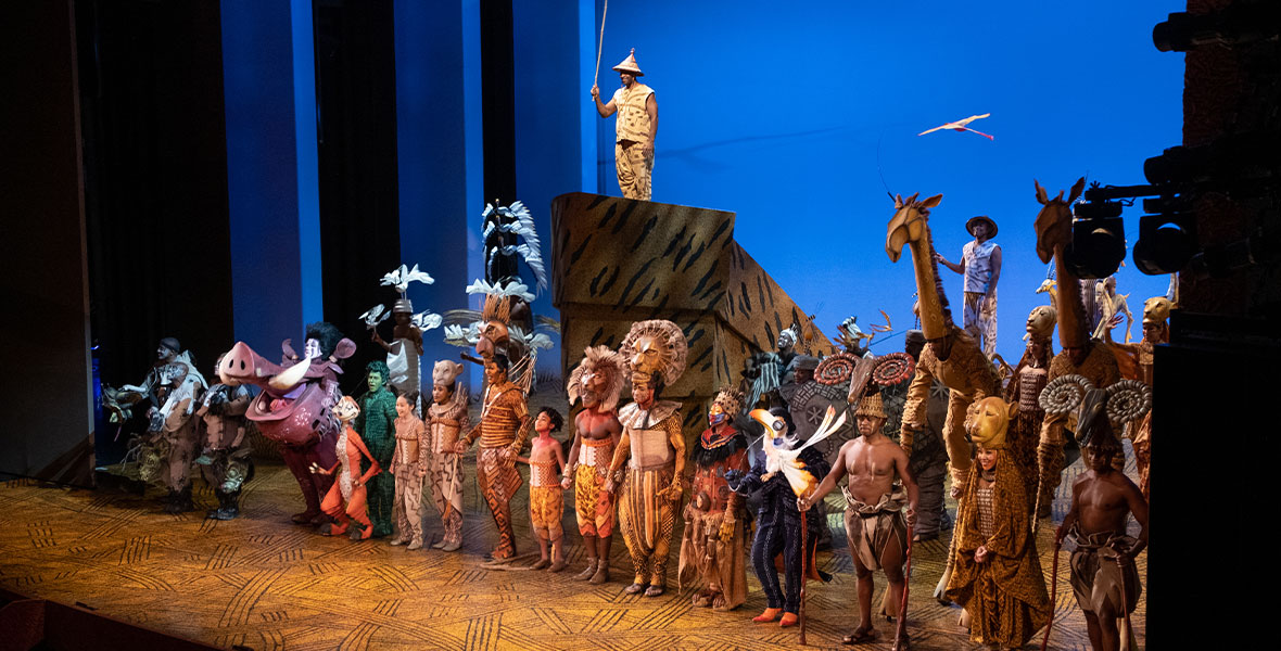In a still from The Lion King on Broadway’s 25th anniversary performance on November 13, 2022, the company is taking their curtain call at the end of the show; they’re holding hands and looking out at the audience, smiling. Almost everyone is standing in a line downstage towards the edge of the physical stage, while one cast member up on “Pride Rock,” holding up a puppet of a bird. The backdrop behind the cast is blue. Photo by Chrystofer Davis.