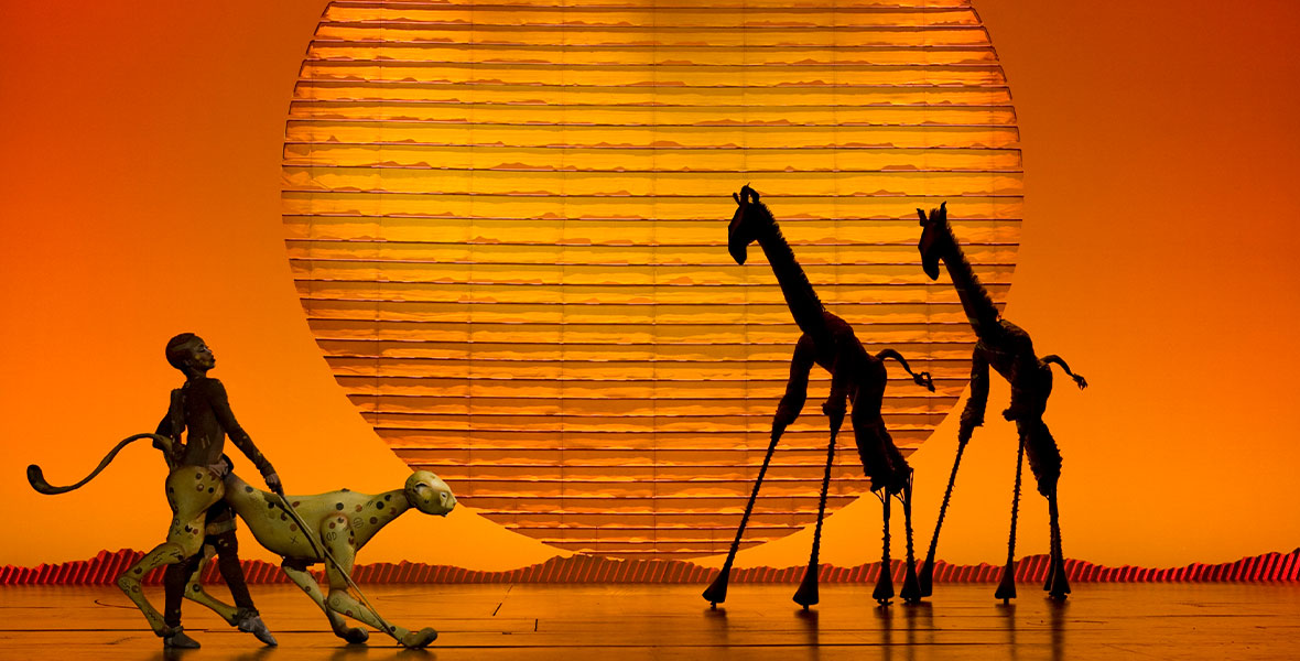 A production still from The Lion King on Broadway, of ensemble members performing as a cheetah and two giraffes, during “Circle of Life.” A large yellow “sun” is seen in the background. Photo by Joan Marcus.