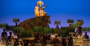 Jayden Theophile as Young Simba and L. Steven Taylor as Mufasa, along with the ensemble of The Lion King on Broadway. Young Simba and Mufasa are up on a set piece, at center, denoting &ldquo;Pride Rock&rdquo;; Mufasa&rsquo;s hand is outstretched. Ensemble members are wearing headdresses outfitted with small platforms of grass. (Photo by Matthew Murphy)