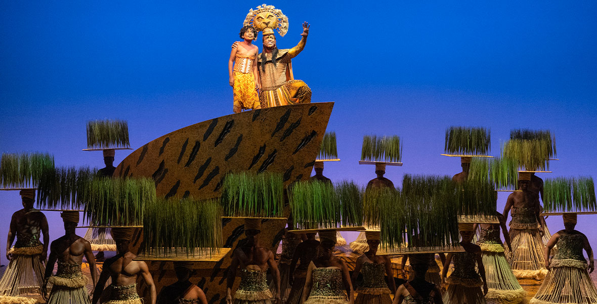 Jayden Theophile as Young Simba and L. Steven Taylor as Mufasa, along with the ensemble of The Lion King on Broadway. Young Simba and Mufasa are up on a set piece, at center, denoting “Pride Rock”; Mufasa’s hand is outstretched. Ensemble members are wearing headdresses outfitted with small platforms of grass. (Photo by Matthew Murphy)