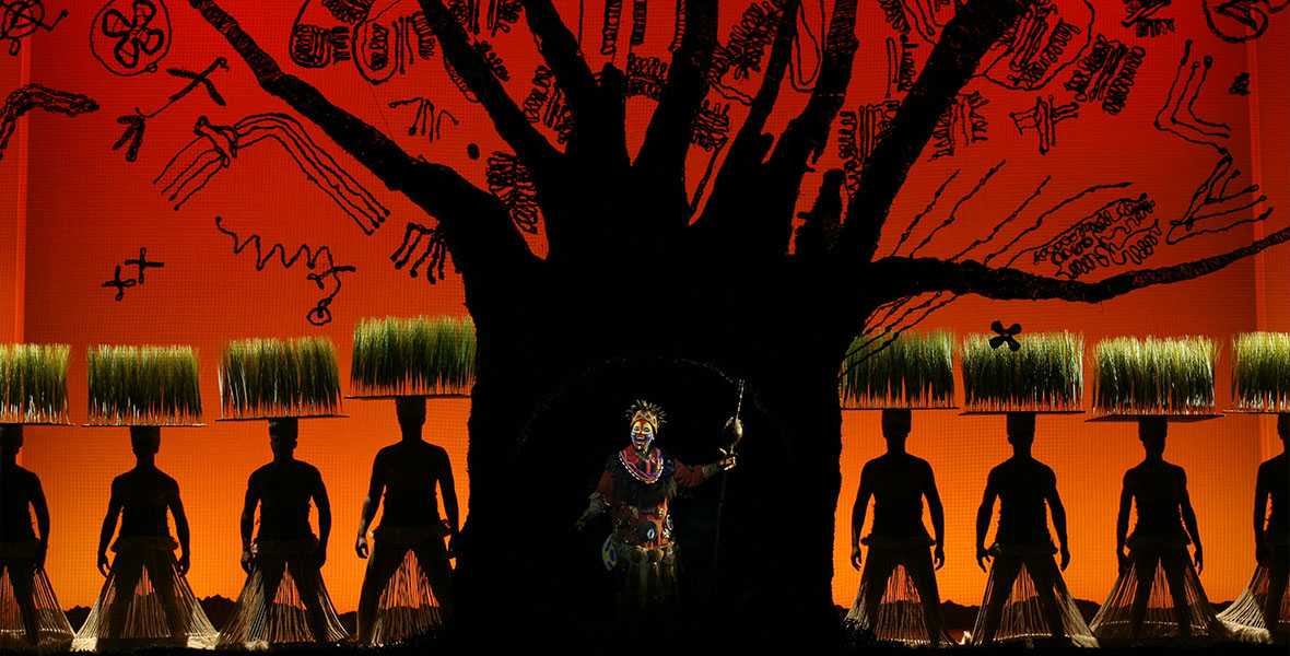 Tshidi Manye as Rafiki and ensemble in a production still from The Lion King on Broadway. Rafiki is standing center, in front of the shadow of a large tree, the branches of which stretch up and surround other hand-drawn images, all set against a reddish orange backdrop. Ensemble members to the left and right of Rafiki are wearing headdresses outfitted with small platforms of grass. (Photo by Joan Marcus)