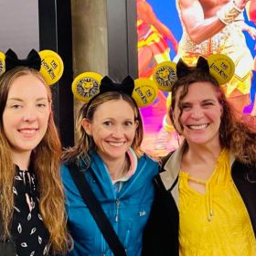 Three fans stand in front of The Minskoff Theatre and The Lion King on Broadway posters. All three fans are wearing Minnie Mouse ears themed to The Lion King. The ears are yellow with The Lion King text and image logo on each side with a black bow in the middle. The guest on the left has long brown hair, a black and white dress, and a black raincoat. The guest in the middle has long blond hair, a blue jacket, and jeans. The guest on the right has long brown hair with a yellow shirt, black jacket, and blue jeans.