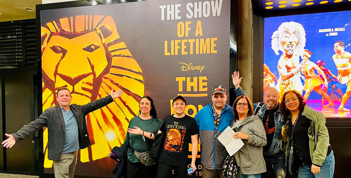 Seven fans stand in front of The Minskoff Theatre and The Lion King on Broadway posters. The fan on the left is wearing a blue shirt, tan slacks, and a black jacket. The next fan to the right is wearing a green shirt with black pants. The next fan to the right has a black baseball cap, black shirt with The Lion King and Simba on it, and black jeans. The fan in the middle is wearing a baseball cap, blue and gray jacket, and gray pants. The next fan to the right has a gray jacket and blue jeans. The next fan to the right has a jean jacket, black shirt, and blue jeans. The fan on the far right has a green jacket, black shirt, and blue jeans. 