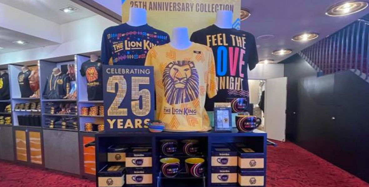 A shelf holds The Lion King 25th Anniversary merchandise. On top of the black shelf are three shirts on mannekins. The shirt on the right is black with The Lion King written in yellow. The shirt in the middle is yellow with The Lion King text and image logo. The shirt on the left is black with “Feel the Love Tonight” written in yellow, blue, and red. Surrounding the shirts are multiple small items. The black shelf has multiple merchandise selections folded and stored.