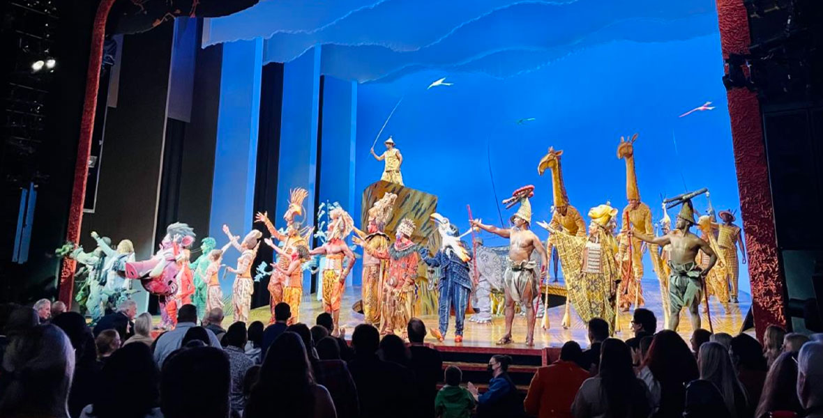 A variety of performers bow on stage. The back of the stage is lit in bright blue. The audience is giving a standing ovation.