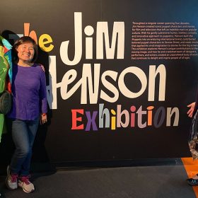 Three guests stand in front of the entrance sign to The Jim Henson Exhibition. The fan on the left has bright red hair with a green flower in it. The fan has green lipstick. The guest is wearing a green dress, dark green leggings, green sneakers, and a light blue jacket with Kermit the Frog on it. The guest has a black purse with a Kermit Numio plush hanging off of it. The guest to the right is wearing a long sleeve purple shirt and blue jeans. The guest on the far left is wearing black glasses, a black shirt, a patterned skirt and purple sweater.