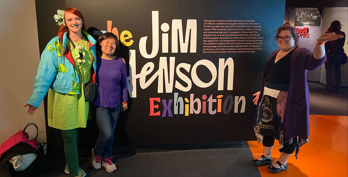 Three guests stand in front of the entrance sign to The Jim Henson Exhibition. The fan on the left has bright red hair with a green flower in it. The fan has green lipstick. The guest is wearing a green dress, dark green leggings, green sneakers, and a light blue jacket with Kermit the Frog on it. The guest has a black purse with a Kermit Numio plush hanging off of it. The guest to the right is wearing a long sleeve purple shirt and blue jeans. The guest on the far left is wearing black glasses, a black shirt, a patterned skirt and purple sweater.