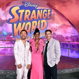 From left to right: Jaboukie Young-White, Gabrielle Union, Jake Gyllenhaal. Young-White smiles and stands in front of a ‘Strange World World Premiere’ sign and platform. He wears a white suit covered in a suit-print pattern. Union smiles and wears a hot pink dress with over-sized rosettes accentuating her neckline. Her hair is styled in a topknot. Gyllenhaal smiles and wears a gray suit jacket over an unbuttoned light pink shirt, which is layered over a black T-shirt. He wears black pants and white sneakers.
