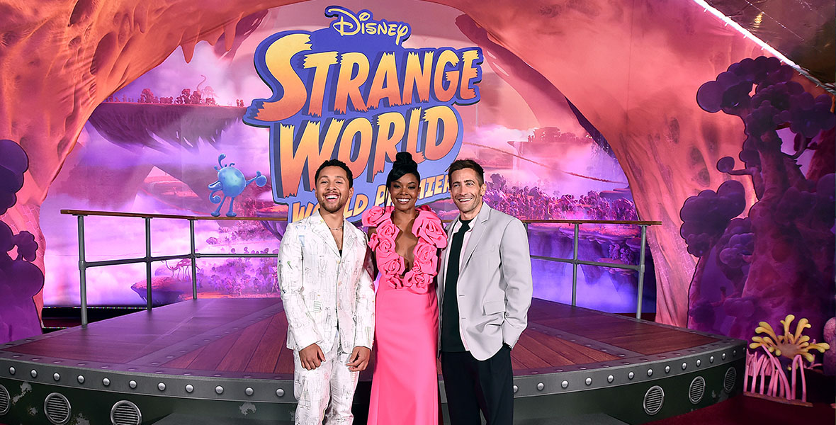 From left to right: Jaboukie Young-White, Gabrielle Union, Jake Gyllenhaal. Young-White smiles and stands in front of a ‘Strange World World Premiere’ sign and platform. He wears a white suit covered in a suit-print pattern. Union smiles and wears a hot pink dress with over-sized rosettes accentuating her neckline. Her hair is styled in a topknot. Gyllenhaal smiles and wears a gray suit jacket over an unbuttoned light pink shirt, which is layered over a black T-shirt. He wears black pants and white sneakers.