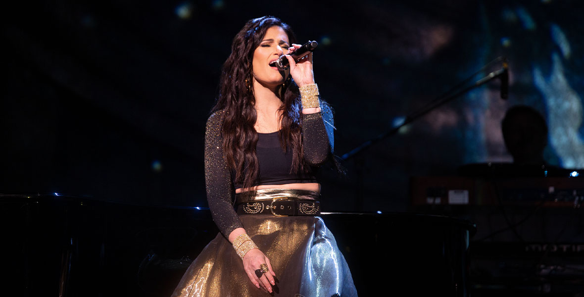 In an image from Idina Menzel: Which Way to the Stage?, Idina Menzel stands center stage and sings into a handheld microphone. She is wearing a gold skirt, a black studded belt, a black crop top with gold sparkles, and gold jewelry.