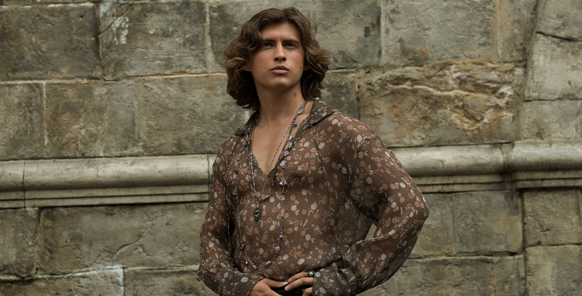 Airk (Dempsey Bryk) stands against a brick wall. He wears a floral, sheer top; layered necklaces; and ornate rings.