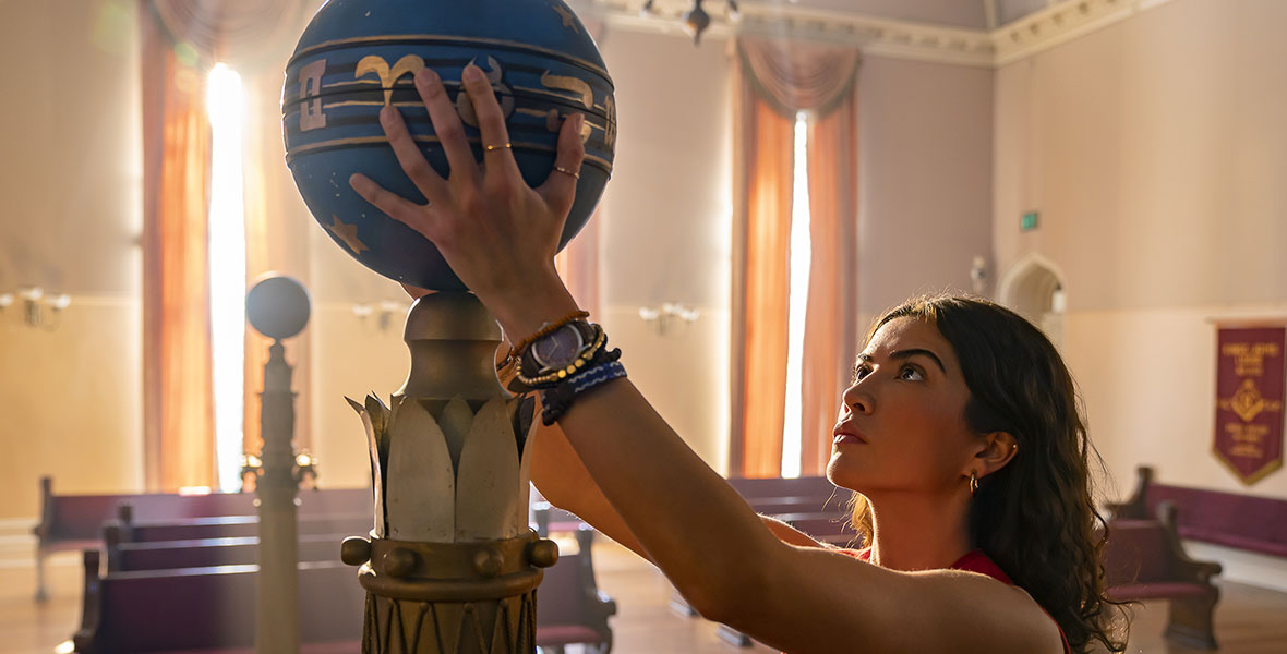 Jess Valenzuela (Lisette Olivera) examines a blue and gold orb in an image from National Treasure: Edge of History. She is wearing a striped tank top and a gold medallion.