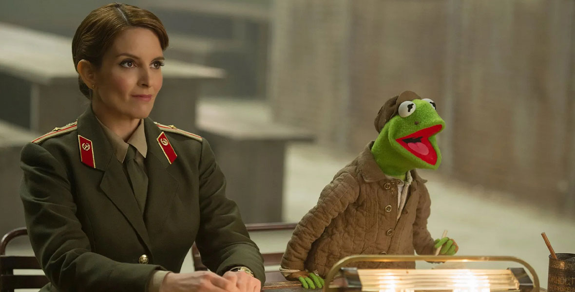Nadya (Tina Fey) wears an olive green blazer with brass buttons, an olive green tie, and a khaki shirt in a still from Muppets Most Wanted. To her left is Constantine, who resembles Kermit the Frog, and wears a matching olive green jacket and hat. They are seated together at a desk.