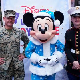 Mickey Mouse, wearing a blue Santa suit with fur trim and a white snowflake print, stands in the middle of two uniformed U.S. Marines. Beside the Marines are decorated Christmas trees. Behind them is a sign that says Disney Ultimate Toy Drive.