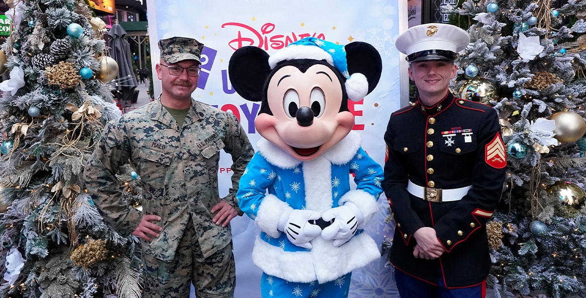 Mickey Mouse, wearing a blue Santa suit with fur trim and a white snowflake print, stands in the middle of two uniformed U.S. Marines. Beside the Marines are decorated Christmas trees. Behind them is a sign that says Disney Ultimate Toy Drive.