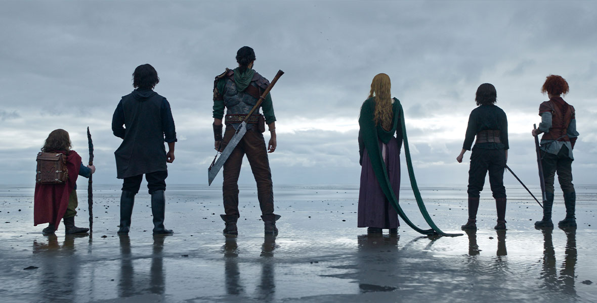 In a scene from Disney+ Original series Willow, the stars of the series stand with their backs toward the viewers as they stare at a large body of water. From left to right stand actors Warwick Davis as Willow Ufgood, Tony Revolori as Graydon, Amar Chadha-Patel as Boorman, Ellie Bamber as Dove, Ruby Cruz as Kit, and Erin Kellyman as Jade. Davis holds a wooden staff in his right hand and wears a maroon cape, olive pants, and brown leather backpack. Revolori wears black knee-high boots, black pants, a black long-sleeved top, and a charcoal hooded vest. Chadha-Patel wears brown boots, brown pants, a green hooded long-sleeved top, and has a large metal weapon slung across his back. Bamber wears a purple skirt and green knitted sweater with two long strands cascading to the wet sand. Cruz wears a dark long-sleeved top, black pants, black knee-high boots, and a leather corset. Cruz holds a sword in her right hand. Kellyman wears olive pants, black knee-high boots, a gray long-sleeved top with a maroon short-sleeved top, and a brown leather vest. Kellyman holds a saber in her left hand.