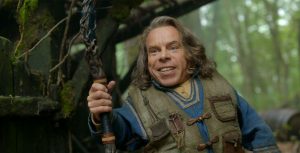 In a scene from Disney+ Original series Willow, actor Warwick Davis portrays Willow Ufgood. He holds a large wooden staff adorned with intricate carved embellishments. He wears a blue, long-sleeved tunic and olive utility vest. He stands in a densely populated forest next to a large tree covered in green moss and vines.