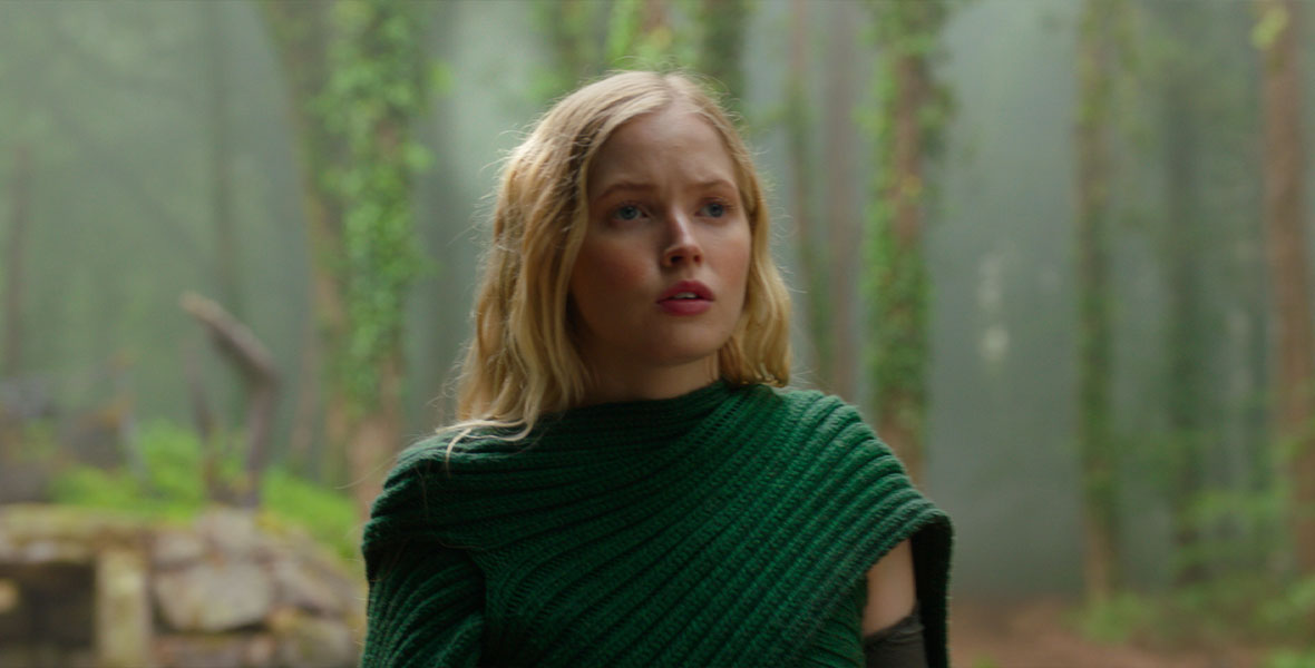 In a scene from Disney+ Original series Willow, actor Ellie Bamber portrays Dove. She wears a green knitted sweater draped across her shoulders. Behind her is a lush, green forest with tall trees covered in moss and ivy.