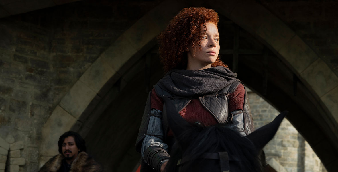 In a scene from Disney+ Original series Willow, Erin Kellyman portrays Jade. She sits atop a black horse and looks to her left as she rides through a stone archway. She wears a maroon top, a gray long-sleeved T-shirt, a black quilted cape, and a black scarf that is draped over her shoulders. Behind Kellyman is actor Tony Revolori as Graydon. He also sits atop a horse and wears a brown fur overcoat.