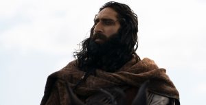 In a scene from Disney+ Original series Willow, actor Amar Chadha-Patel portrays Boorman. He sits atop a large black horse and looks to his right. He wears a tan long-sleeved top, brown overcoat, and matching brown scarf draped across his shoulders. Behind him is a blurred gray and light blue sky.