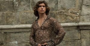 In a scene from Disney+ Original series Willow, actor Dempsey Bryk portrays Airk. He stands in front of a stone wall with his hands resting on his left hip. He wears a sheer brown blouse with peach and tan flowers, black pants with white stripes, and silver necklaces and rings.