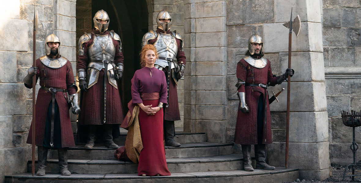 In a scene from Disney+ Original series Willow, actor Joanne Whalley portrays Sorsha. She stands on stone steps and surrounded by four men wearing metal armor and maroon tunics. She wears a purple blouse and a cardinal maxi skirt with maroon and gold fabric wrapping her waist and draping down her side. They stand in front of a tall stone archway with a gravel pathway in front of them.