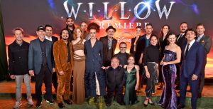 The cast and crew of Willow on the green carpet at the series’ world premiere. Nearly two dozen crew members stand smiling against a large Willow Premiere backdrop, with Warwick Davis front and center in a black suit.