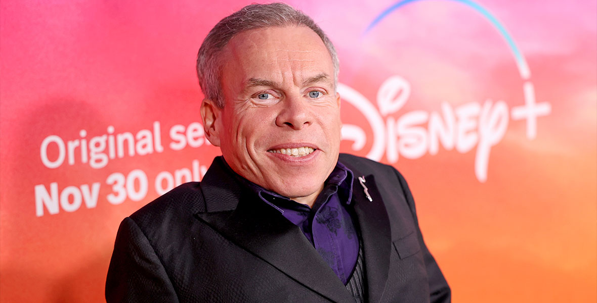 Warwick Davis smiles at the camera and stands against a bright pink and orange backdrop. He is wearing a black suit jacket with a purple shirt underneath.