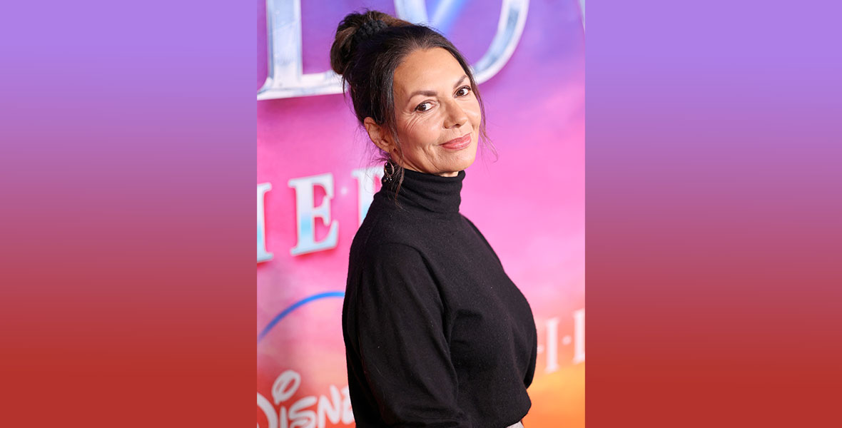 Joanne Whalley gives a tight slipped smile over her shoulder as she stands against a bright pink and purple backdrop. She has her dark brown hair up in a bun and is wearing a soft, black turtleneck.