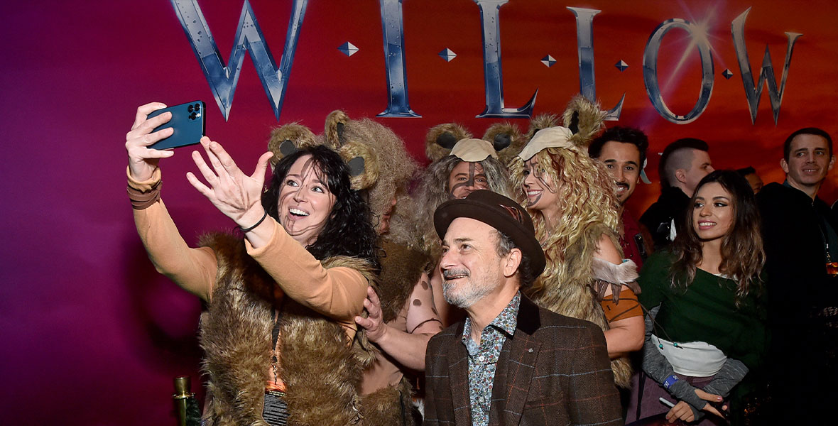 Fans dressed in elaborate Willow costumes crowd around actor Kevin Pollak for a selfie. Kevin is wearing a brown plaid 3-piece suit and matching brown fedora.
