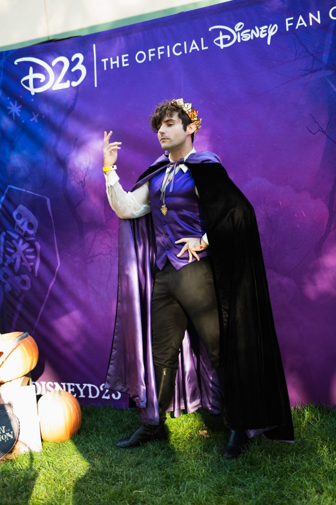 A guest posing as the evil queen in front of the D23 photo opportunity, which has a blue and purple background with “D23: The Official Disney Fan Club” in white above and spooky designs faded in the back. The guest posing has a black velvet cape with a purple lining, a deep purple vest, long white sleeve shirt, black pants, and black shoes, as well as gold jewelry with a gold and red crown.
