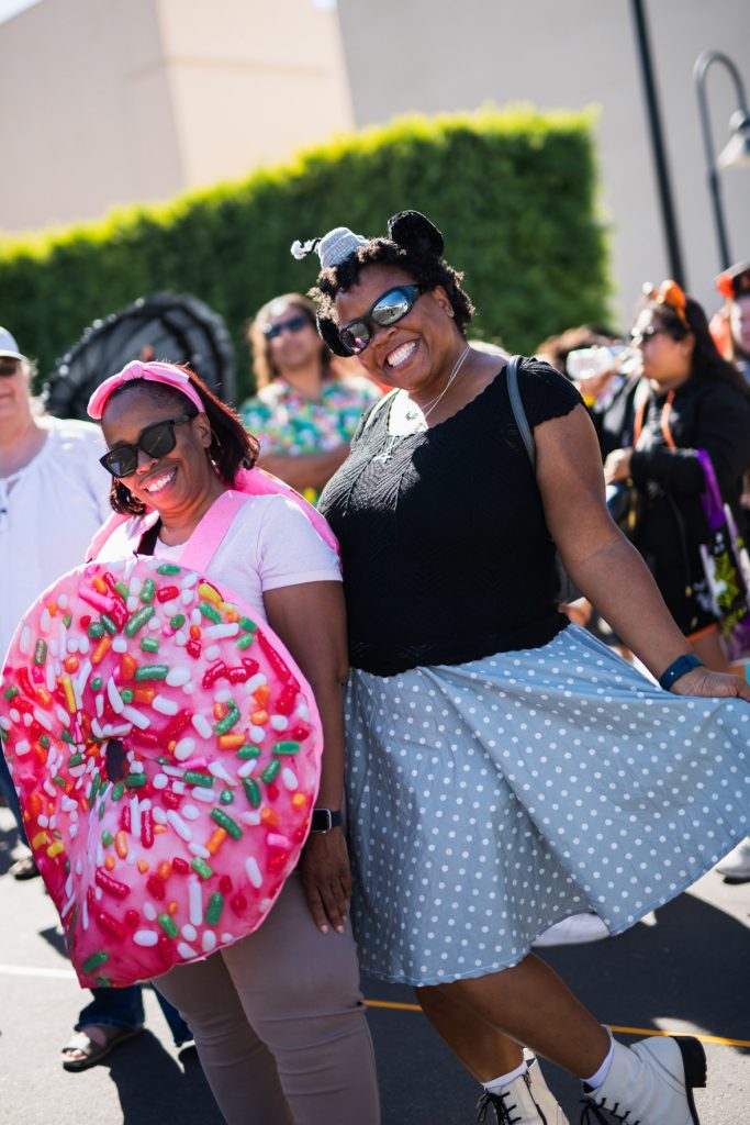 Two guests dressed as a donut and Minnie Mouse. The guest on the left has a white shirt with brown pants and a pink sprinkled donut pull-over. The guest on the right has a small grey hat attached with Minnie ears, black shirt, and grey and white polka dot skirt.