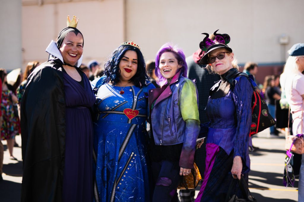 Four guests dressed as the Evil Queen, Evie, Mal, and Maleficent from Descendants. The guest on the right is dressed as the Evil Queen with a long purple dress, black cape and gold crown. The guest to their right is dressed as Evie with blue curled hair, gold crown, and royal blue dress with gold and red detailing. The guest to their right is dressed as Mal with purple hair, purple and green leather jacket, and purple pants. The guest to their right is dressed as Maleficent with Maleficent Minnie ears, purple top and black skirt.